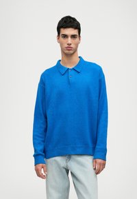 Maglione blu a maglia con colletto, caratterizzato da maniche lunghe, una vestibilità comoda e un orlo a coste. Un paio di jeans blu chiaro completa il look.