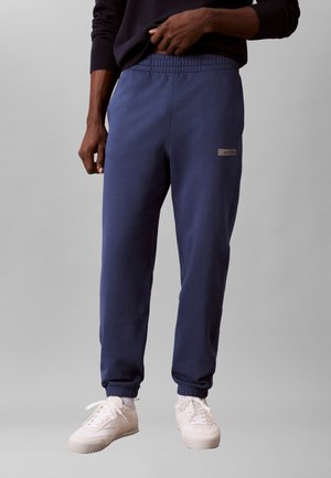 Mand iført marineblå Calvin Klein joggers med elastiske manchetter og hvide sneakers, stående mod en ensfarvet grå baggrund.