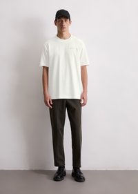 White cotton t-shirt with short sleeves, paired with dark olive green tailored trousers and black lace-up shoes. Black cap worn on head.