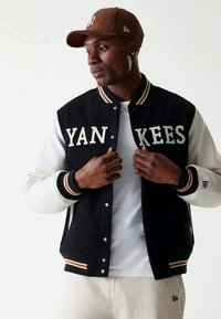 Moški z rjavim baseball klobukom in mornarsko modrim ter belim Yankees jakno, z belim majico in svetlim hlačami, prilagaja sprednji del jakne.