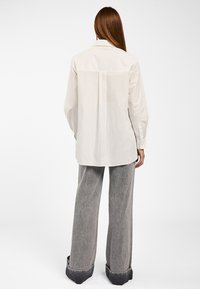 Weißes Langarmhemd mit einem Rückenfalten, aus glattem Material. Kombiniert mit weiten grauen Jeanshosen mit rohen Säumen und kontrastierenden Bündchen.