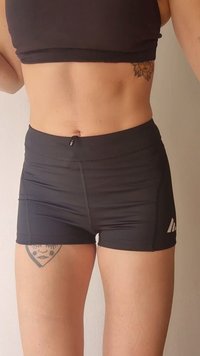 Person trägt schwarze Sportshorts und ein schwarzes Croptop, zeigt Tätowierungen am linken Oberschenkel und an der rechten Seite des Oberkörpers.