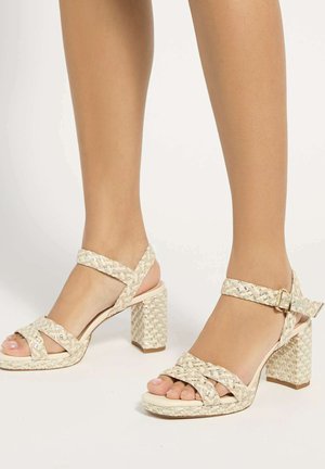 Pieds portant des sandales à talons blocs tissées beiges avec des brides à la cheville et des boucles dorées sur un fond blanc.