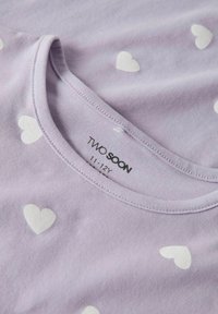 T-shirt lilas avec motif de cœurs blancs, fabriqué en tissu doux. Il a un col rond et une étiquette imprimée indiquant "TROP TÔT" et la taille.