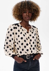 Beżowa bluzka z długim rękawem w czarne kropki, z zapięciem na guziki z przodu i koronkowymi akcentami na ramionach. Połączona z ciemnymi dżinsami.
