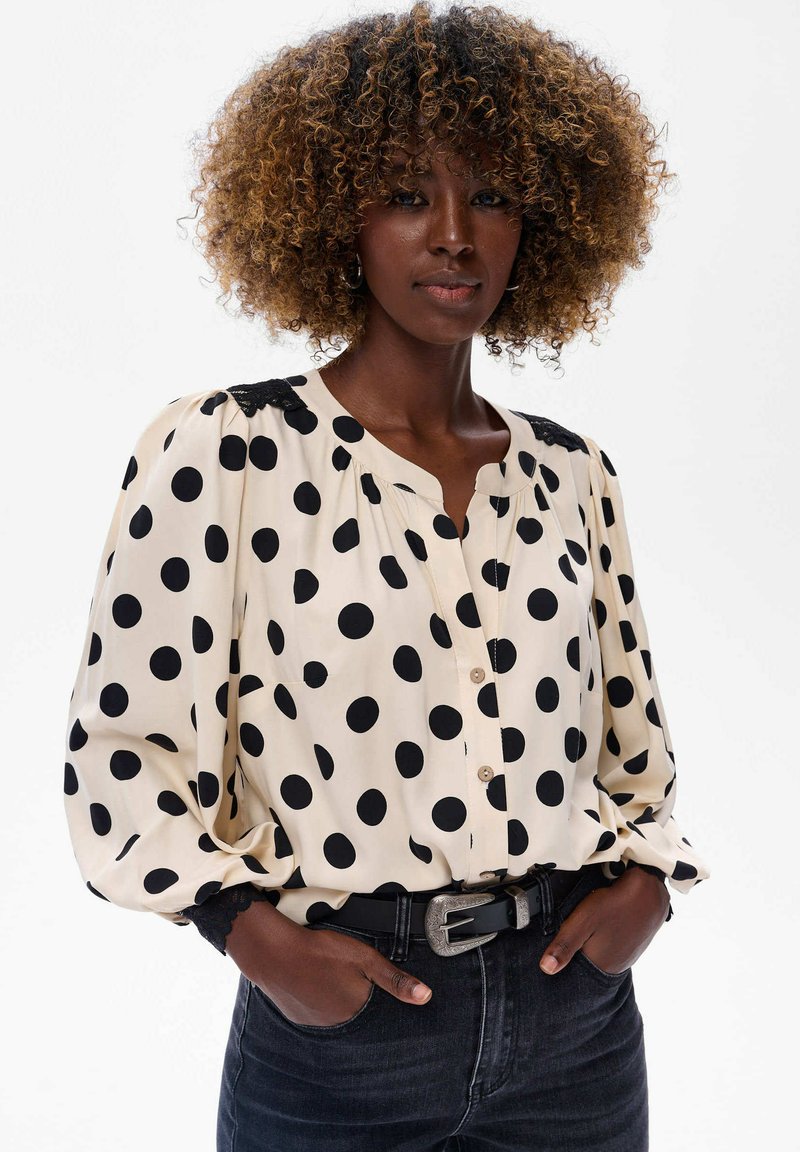 Beżowa bluzka z długim rękawem w czarne kropki, z zapięciem na guziki z przodu i koronkowymi akcentami na ramionach. Połączona z ciemnymi dżinsami.