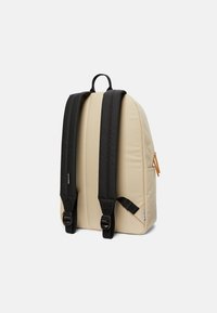 Zaino beige con spallacci neri ergonomici, manico superiore, tasche laterali e una cerniera di colore beige; materiale liscio e imbottito per il massimo comfort.