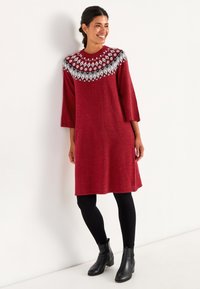 Rød strikket kjole med et mønstret rundt halsudskæring i sort og hvid, trekvartærmer og en afslappet pasform, parret med sorte leggins og ankelstøvler.