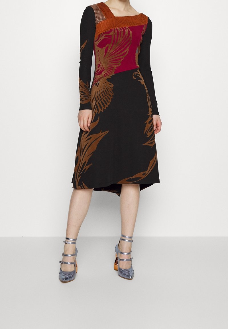 Femme portant une robe noire avec des motifs d'oiseaux et de feuilles marron et rouges, manches longues, encolure carrée et chaussures à talons hauts argentées à brides.