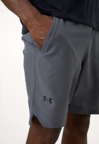 Pilkos sportinės šortai su tekstūruota medžiaga, turintys šonines kišenes ir juodą "Under Armour" logotipą kairėje apatinėje pusėje.