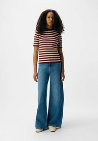 Haut à manches courtes rayé en bordeaux et crème, associé à un jean bleu à jambes larges ; le mannequin se tient droit avec les mains sur les côtés.