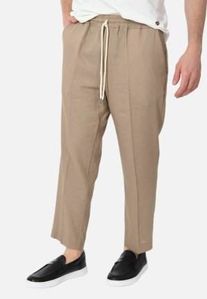 Uomo con pantaloni beige con coulisse e scarpe slip-on nere, in piedi con le mani rilassate lungo i fianchi.