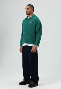 Groene fleece trui met kraag en geborduurd logo, gecombineerd met donkere wijde jeans en zwarte schoenen met contraststiksels.