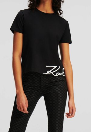 Vrouw draagt een zwart T-shirt met korte mouwen met witte sierlijke tekst bij de zoom en nauwsluitende zwarte broek met patroon.