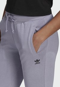 Ruka v kapse světle fialových Adidas tepláků s šňůrkou v pase, nošeno s černým topem.