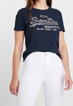 Marineblaues Baumwoll-T-Shirt mit Rundhalsausschnitt und kurzen Ärmeln, versehen mit silbernem Glitzerschriftzug und legerer Passform, kombiniert mit weißen Jeans.