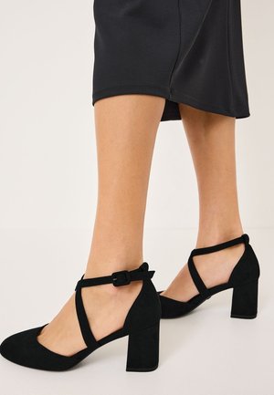 Sort ruskind sko med lukket tå og blokhæl, krydsede ankelremme og en sidespænde på hvid baggrund.