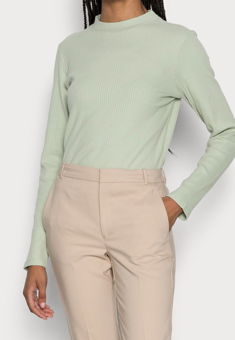 Maglietta a maniche lunghe a coste verde chiaro abbinata a pantaloni beige su misura. Le caratteristiche principali includono un collo alto e una silhouette aderente.