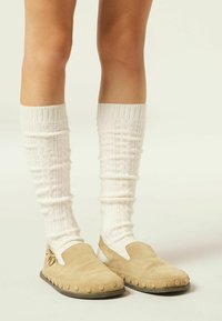Beżowe zamszowe buty slip-on z zaokrąglonym czubkiem i okrągłymi akcentami wzdłuż podeszwy, zestawione z białymi teksturowanymi skarpetami do kolan.