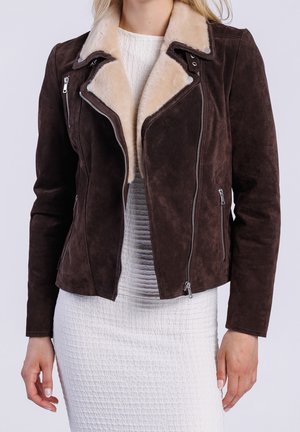 Woman wearing dark brown suede jacket with beige shearling collar over textured white dress, standing with arms relaxed by sides.