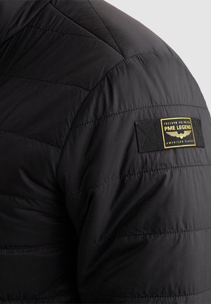 Black quilted jacket sleeve with stitched PME Legend patch in yellow and black displaying winged emblem and text.