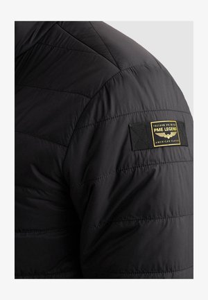 Manche de veste matelassée noire avec un patch PME Legend cousu en jaune et noir affichant un emblème ailé et du texte.