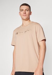 Helles beiges T-Shirt mit kurzen Ärmeln aus Baumwolle. Verfügt über ein gesticktes Logo "Champion" in einer dunkleren Nuance, mit einem kleinen Logo auf dem linken Ärmel.
