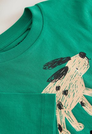 Camiseta de algodón verde con cuello redondo acanalado, que presenta una ilustración de un perro en beige y negro en la parte frontal. Textura suave, mangas cortas.