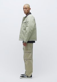 Ανοιχτό πράσινο, καπιτονέ μπουφάν με καφέ γιακά, συνδυασμένο με μπεζ cargo παντελόνι που διαθέτει πολλές τσέπες, και μαύρα αθλητικά παπούτσια.