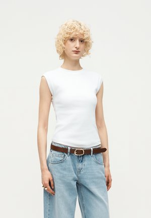 Personne aux cheveux blonds bouclés portant un haut blanc sans manches, un jean bleu clair, une ceinture marron et une bague à la main gauche sur un fond uni.