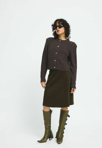 Mörkbrun stickad cardigan med kabelmönster, silvriga knappar, olivgrön kordflöjelkjol och knähöga gröna stövlar med spännen.