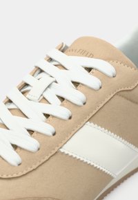 Μπεζ αθλητικό παπούτσι με απαλό σουέντ φινίρισμα, λευκά κορδόνια και λευκή πλαϊνή ρίγα. Διαθέτει στρογγυλεμένη μύτη και υφασμένο καουτσούκ πάτο.