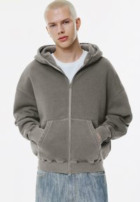 Gray zip-up hoodie made of cotton, featuring a large hood, front pockets, and ribbed cuffs. Paired with light blue denim jeans.