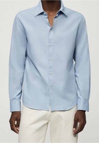 Chemise bleu clair à manches longues en tissu à texture subtile, dotée d'un col pointu et d'une fermeture boutonnée sur le devant, portée avec un pantalon beige.