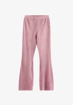 Ung kvinde med krøllet hår iført en navy cardigan, blomster-top, lyserøde bukser og hvide sneakers, som står og smiler foran en ensartet baggrund.