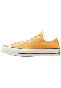 Gule lerret-sneakers med hvite snøring, gummikappe, og svarte detaljer på sålen. Lågtoppsdesign med teksturert overdel.