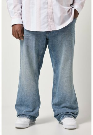 Man draagt losse lichtblauwe spijkerbroek en witte sneakers, met één hand in zijn zak en de andere ontspannen langs zijn zijde.