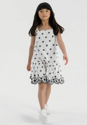 Robe blanche avec broderie florale noire et ourlet en dentelle. Présente un décolleté carré et un bas à volants en couches. Associée à des baskets blanches.