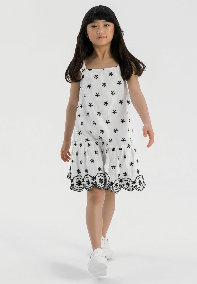 Robe blanche avec broderie florale noire et ourlet en dentelle. Présente un décolleté carré et un bas à volants en couches. Associée à des baskets blanches.