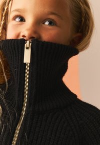 Maglione nero a coste con colletto alto, dotato di dettaglio con zip dorata sul davanti. La trama morbida accentua calore e comfort.
