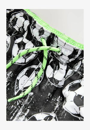 Peldbikses ar melna un balta futbola bumbas rakstu, elastīgu jostasvītu ar zaļu apdari un zaļu saites regulēšanai.