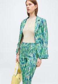 Γυναίκα που φοράει ένα κοστούμι με πράσινο και μπλε μοτίβο, με μπεζ ριμπ τοπ, κρατώντας μια ανοιχτό κίτρινη τσάντα σε λευκό απλό φόντο.