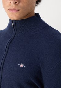 Marinblå zip-up-tröja i mjukt material. Hög krage, subtil textur och broderad logotyp på bröstet. Silverfärgad dragkedja som accent.