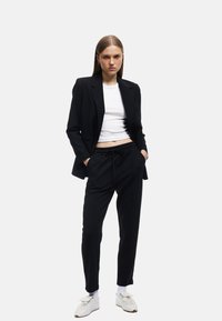 Zwarte blazer over een witte cropped top, gecombineerd met zwarte joggerbroeken. Het model draagt witte sneakers en toont een moderne, ontspannen uitstraling.