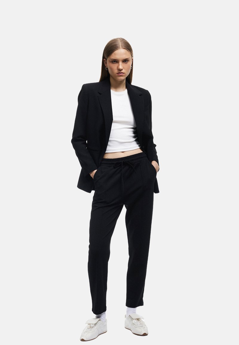 Zwarte blazer over een witte cropped top, gecombineerd met zwarte joggerbroeken. Het model draagt witte sneakers en toont een moderne, ontspannen uitstraling.