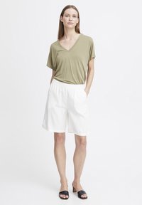 T-shirt vert olive en V et shorts blancs à taille élastique ; design décontracté avec une texture lisse, complété par des sandales à talons noires.