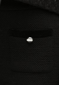 Gros plan d'une poche en tissu noir texturé avec un bord en velours noir et un petit bouton argenté gravé "maje".