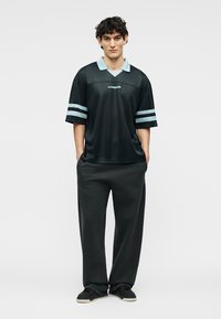 Jeune homme debout, mains dans les poches, portant une chemise noire avec un col et des rayures bleu clair, un pantalon large noir et des baskets noires.