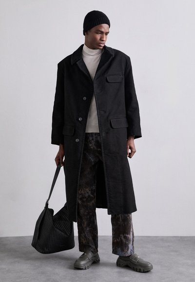 Black long coat with a notched collar, button closure, and front pockets. Worn over a light turtleneck and marbled patterned pants. Holds a textured bag.