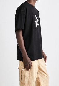 Czarny, oversize t-shirt z graficznym nadrukiem ptaków z przodu. Noszony z beżowymi spodniami cargo z delikatnym wzorem i widocznymi kieszeniami.
